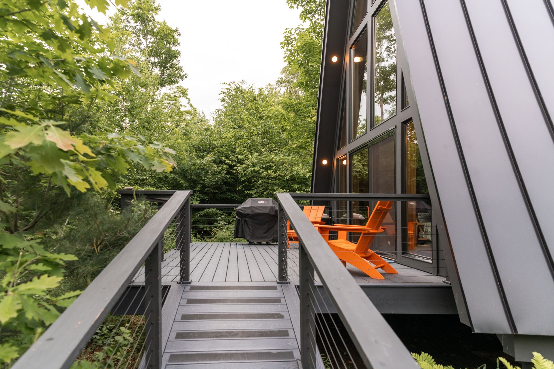 A modern A-frame cabin surrounded by lush greenery with an outdoor deck featuring bright orange chairs.